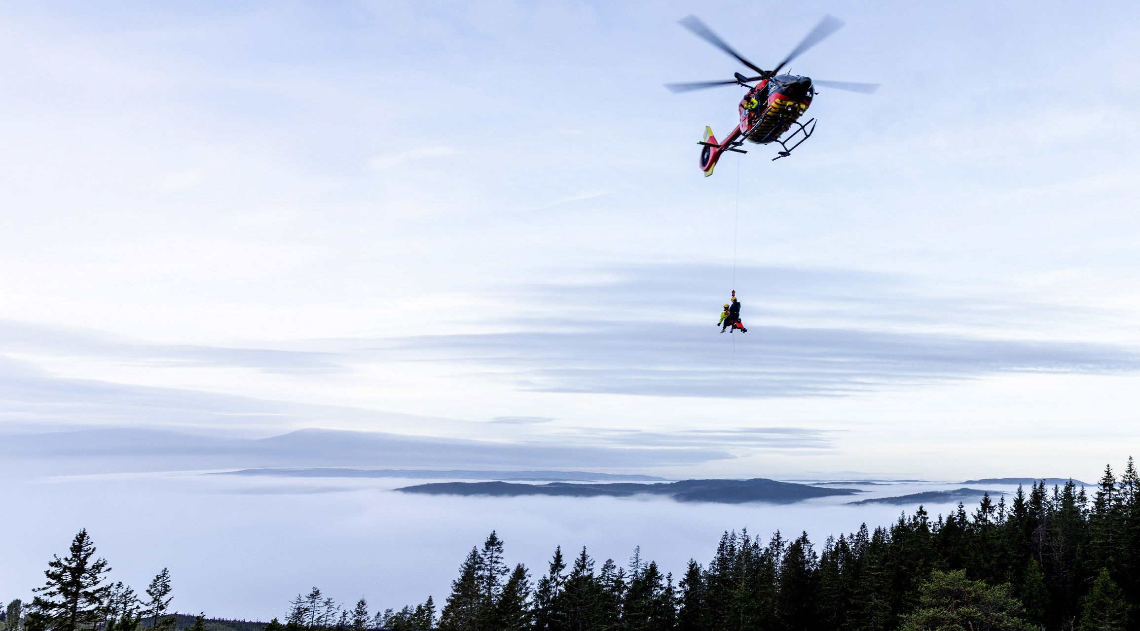 Rescuer on a hoist line above a treeline with a cloud sea below